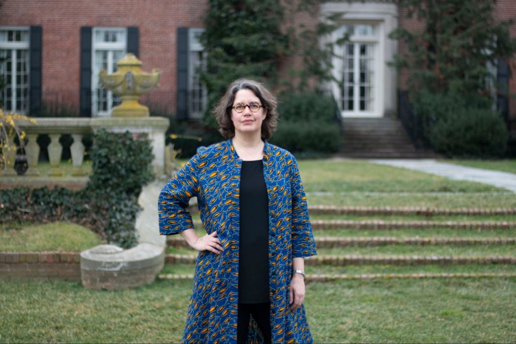 Picture of woman wearing long blue coat with a black shirt. Has Short brown hair and wearing glasses