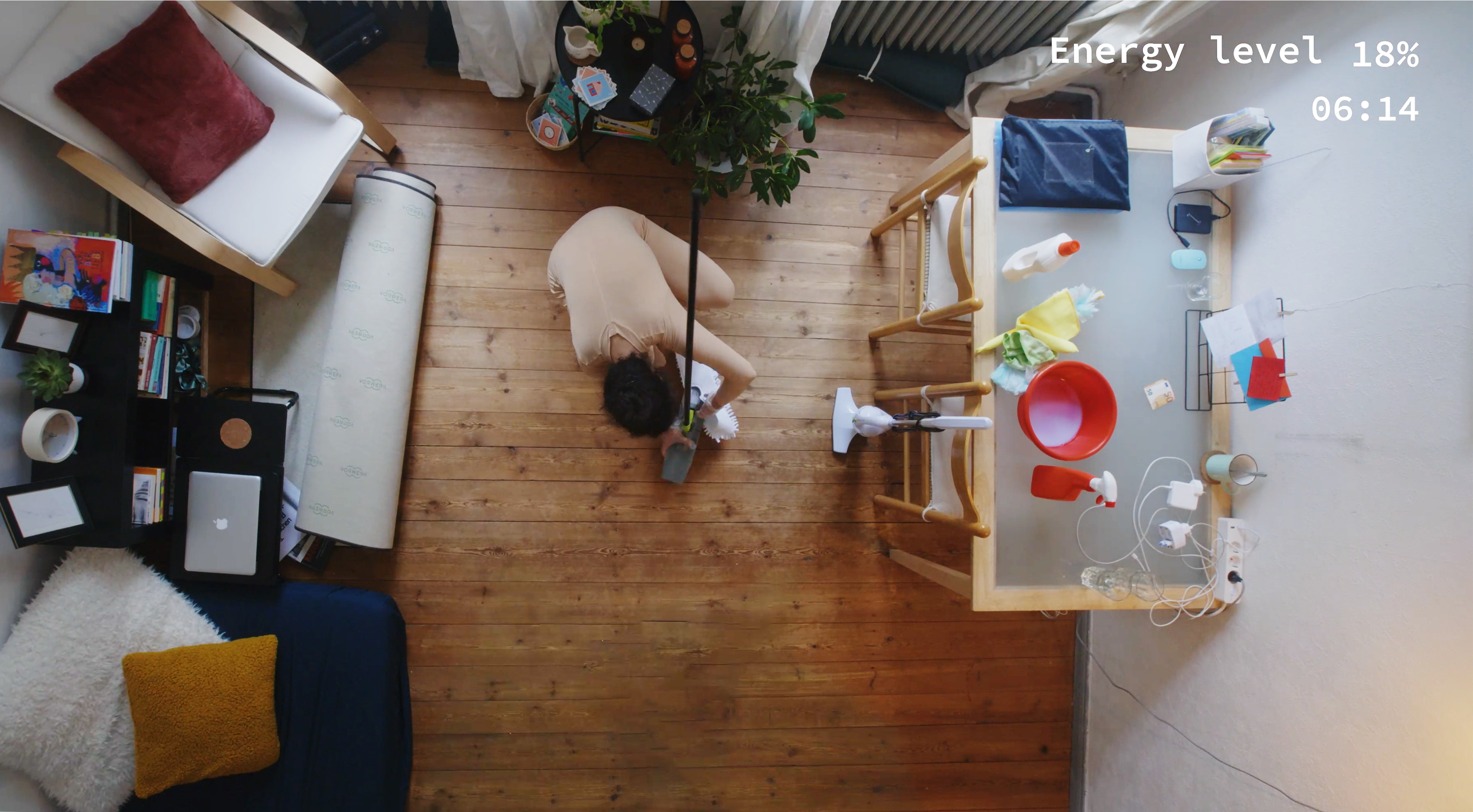 bird's eye view of a woman cleaning a living room, she is crouching on the floor with a mop, other cleaning equipment is visible on a table, text reads: 'energy level 18%, 06:14'
