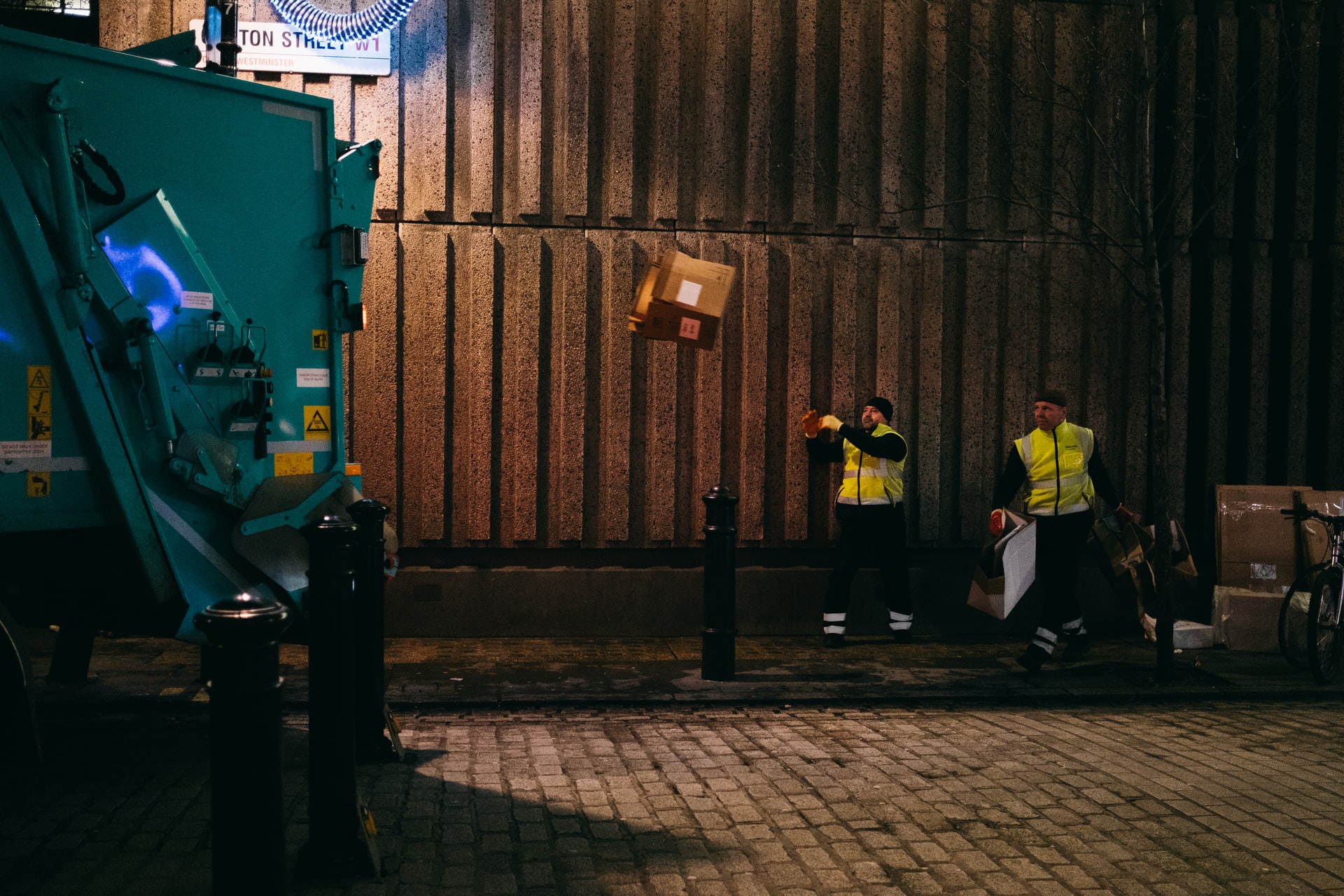 photograph of 2 men wearing hi-viz jackets and throwing empty cardboard boxes onto a rubbish collection truck at night-time