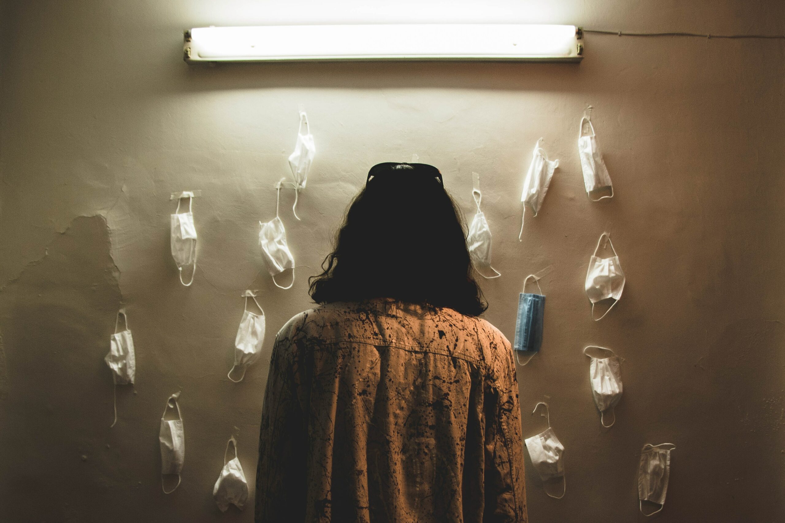 photograph of the back of a woman stnaidng in front of a wall where several facemasks hang